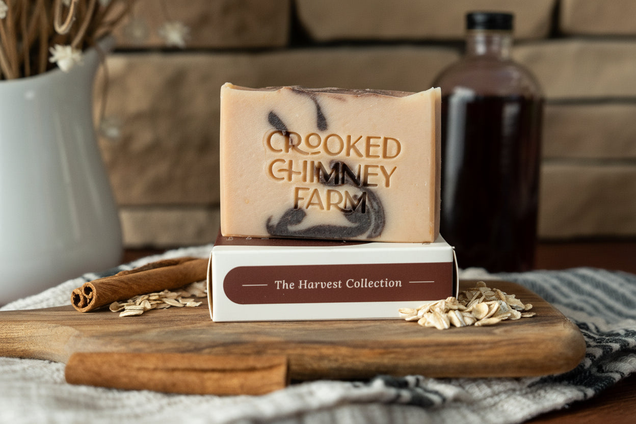 A handmade bar of soap from Crooked Chimney Farm sits on top of its branded box, styled on a wooden board with cinnamon sticks, oats, and a dark bottle in the background.